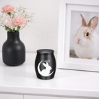 De laatste aai mini urn voor konijn van matzwart RVS met wit silhouet van een konijntje in de maan op een witte kast.