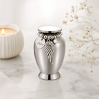 De laatste aai mini urn van RVS met gepersonaliseerd naamplaatje, engelenvleugel bedel en steentje op marmeren tafel.