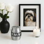 De laatste aai zilverkleurige RVS mini urn met hartjesmotief, gegraveerd met naam, naast een fotolijst en kaars.